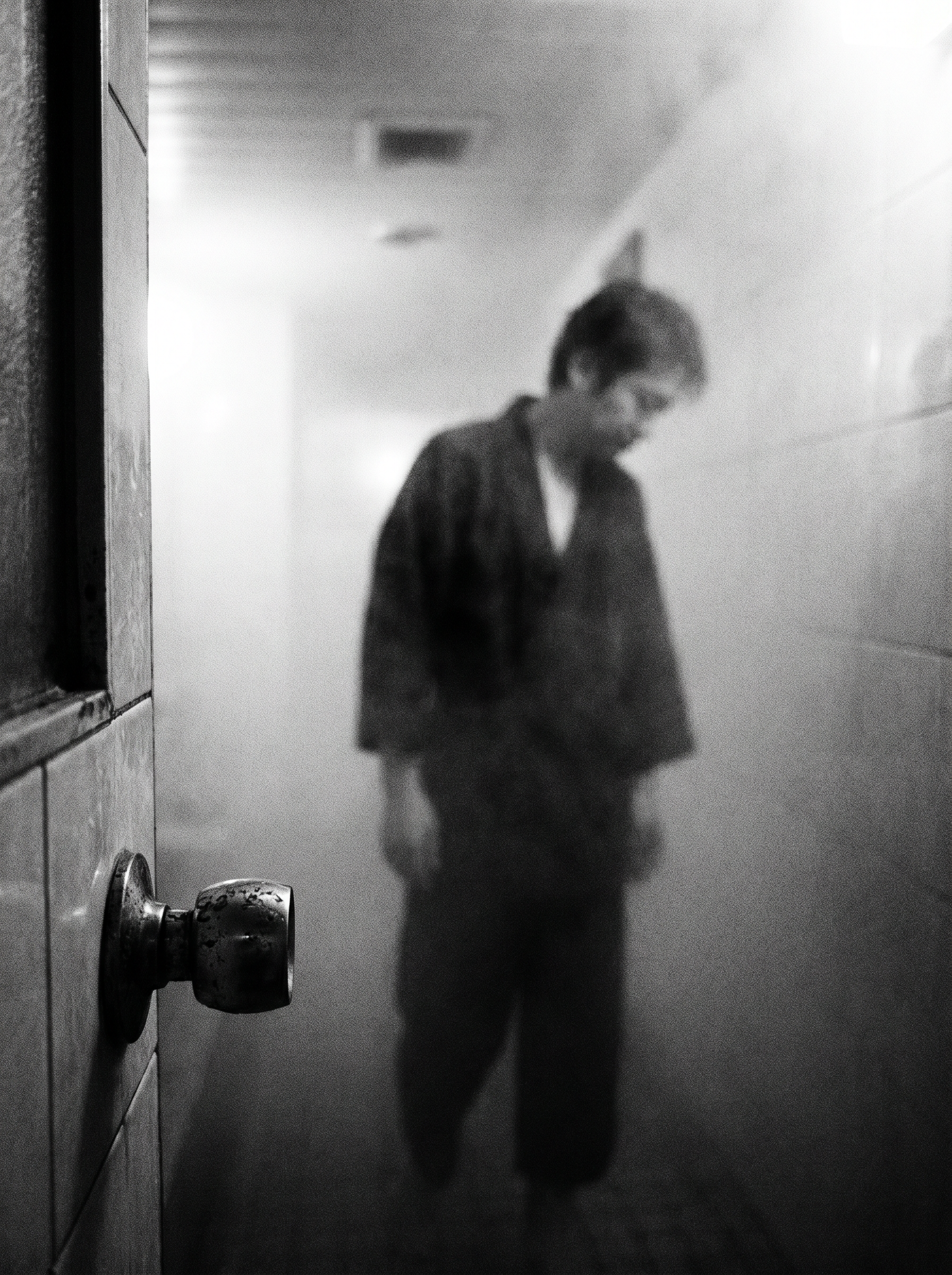 A figure absorbed in dense steam. Head down. Passive. The only sharp element in the frame is the door handle — unused. Fuji X-Pro1 aesthetic, Saul Leiter and Daido Moriyama lineage.